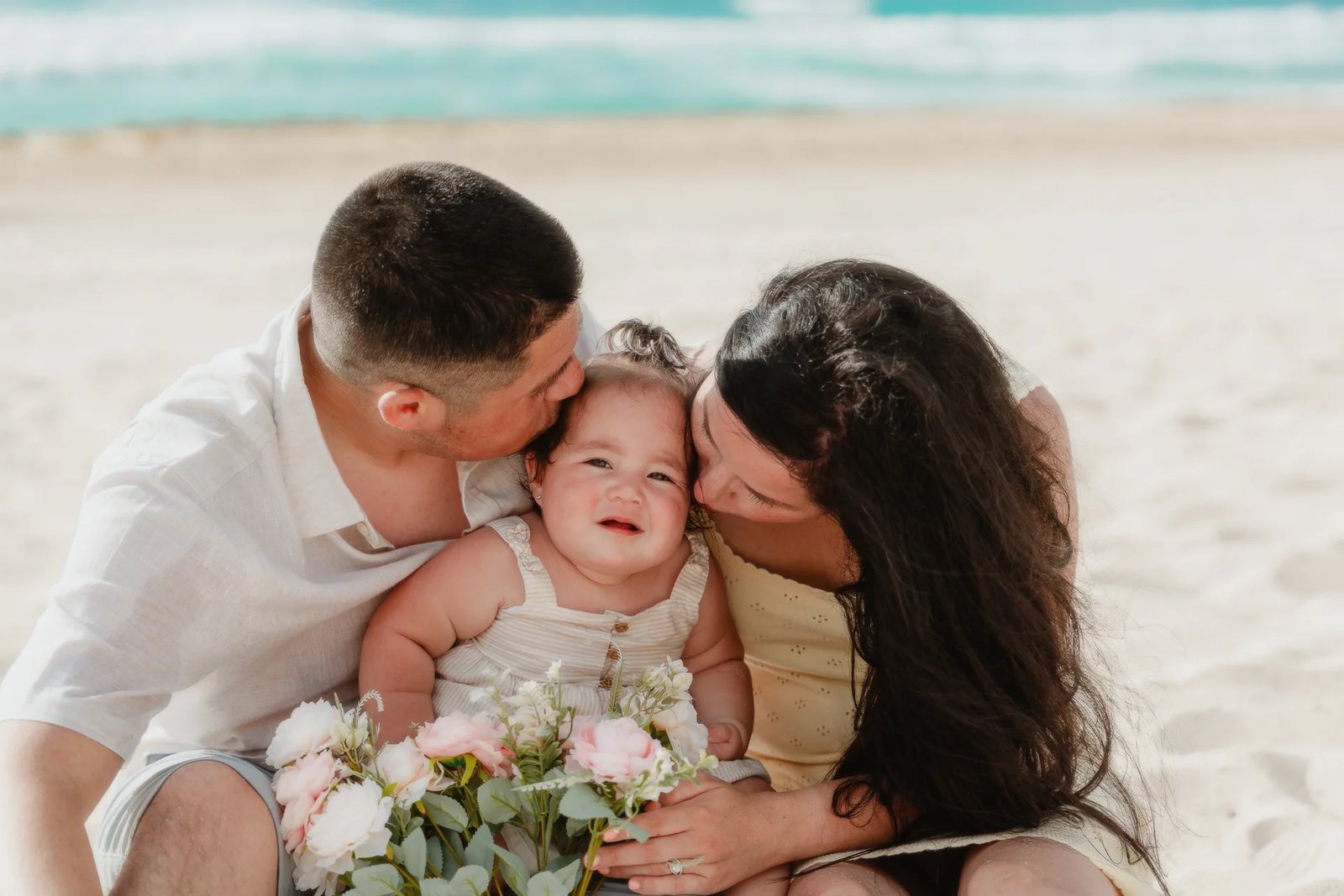 Parents kissing their baby on the sand, with flowers