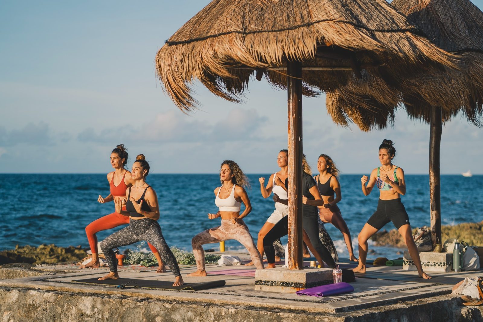A group in a standing yoga pose under a palapa by the sea