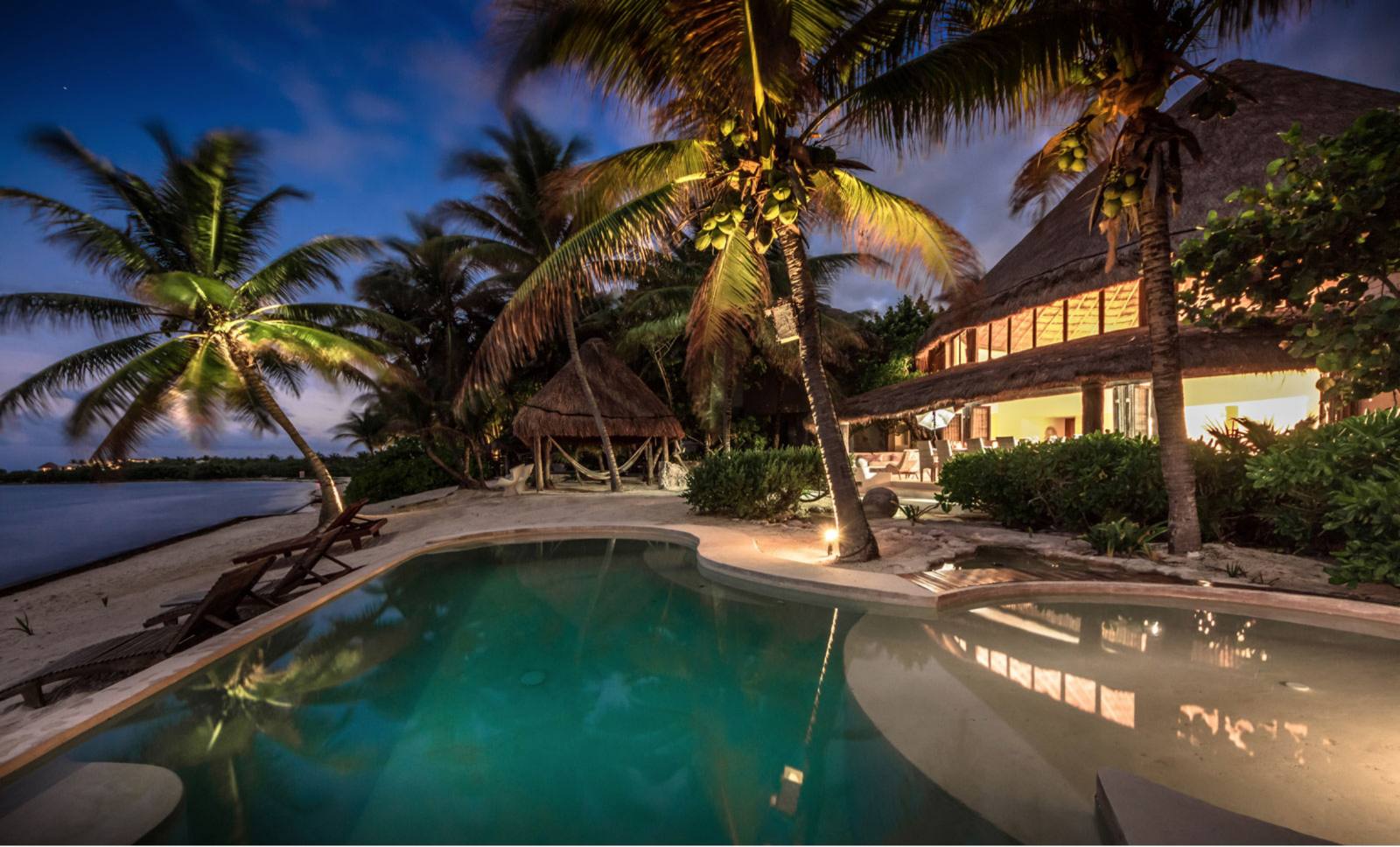 The pool lit at night, with the palapa house behind